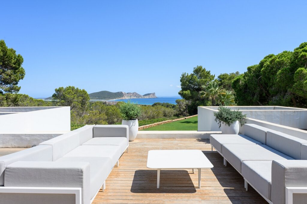 Spacious outdoor terrace with modern white sofas and a table on wooden decking, overlooking lush green trees and a scenic view of the sea and distant hills under a clear blue sky. David Pearson Travel