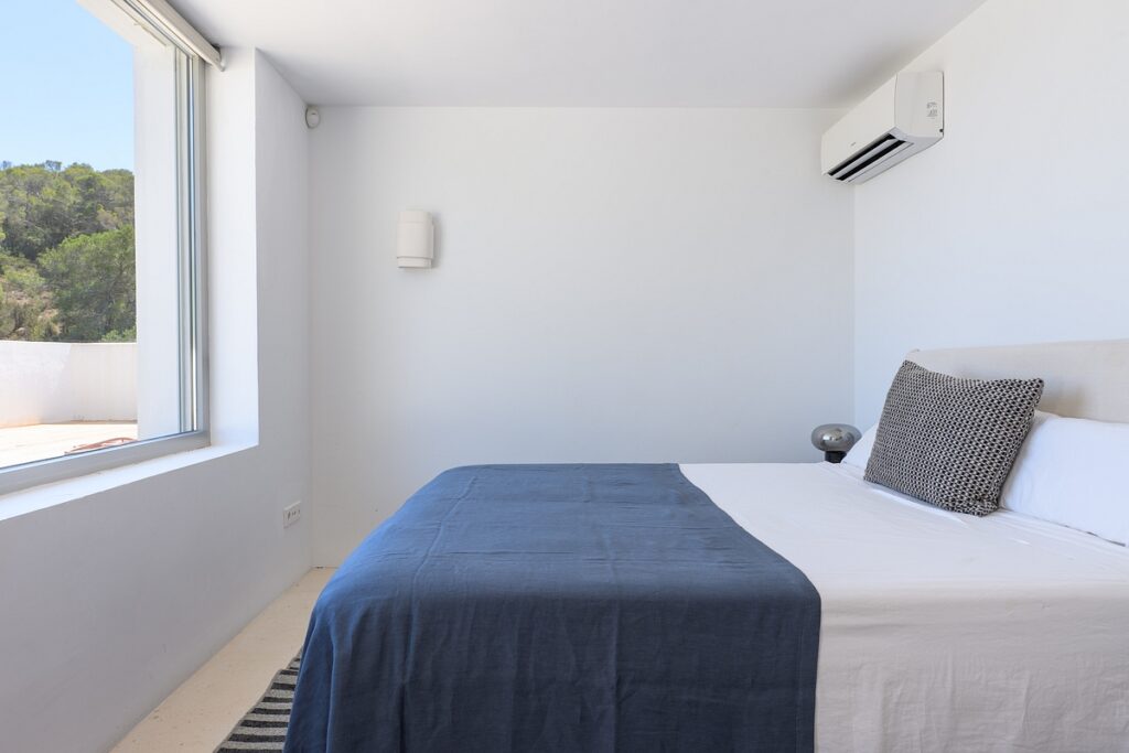 Minimalist bedroom with a large window, white walls, a bed with white linens and a dark blue throw, a patterned pillow, a striped rug, and a wall-mounted air conditioner. Trees are visible outside the window. David Pearson Travel