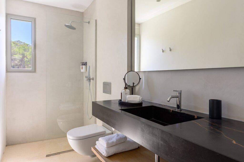 Minimalist bathroom with a walk-in glass shower, wall-mounted toilet, black countertop sink, round mirror, towels, and a large horizontal mirror above the vanity. Natural light comes in from a window by the shower. David Pearson Travel