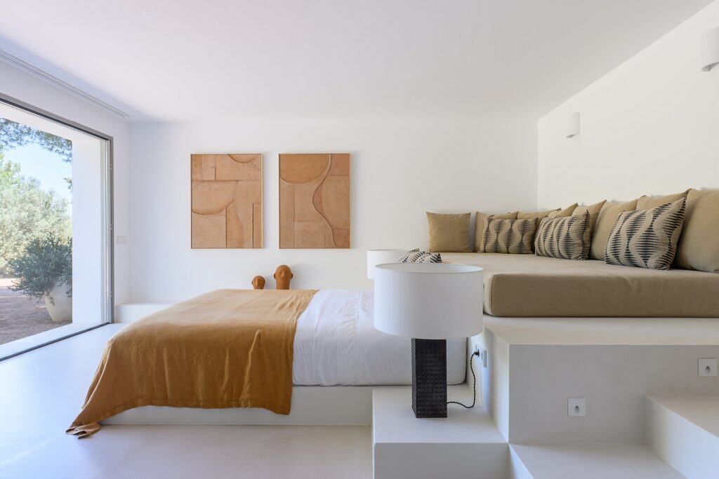 Modern minimalist bedroom with a large window, bed with a mustard blanket, built-in sofa with patterned cushions, abstract wall art, and neutral tones throughout. Natural light fills the room. David Pearson Travel