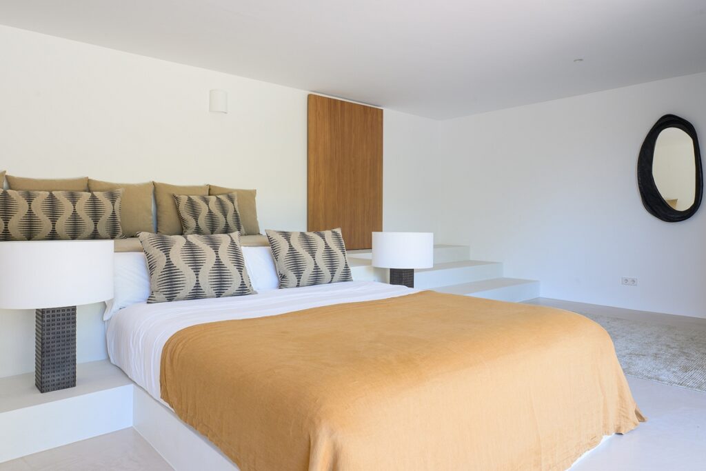 A modern, minimalist bedroom with a low platform bed, mustard-colored bedding, geometric-patterned pillows, two bedside lamps, white walls, a wooden panel, and an oval mirror on the wall. David Pearson Travel