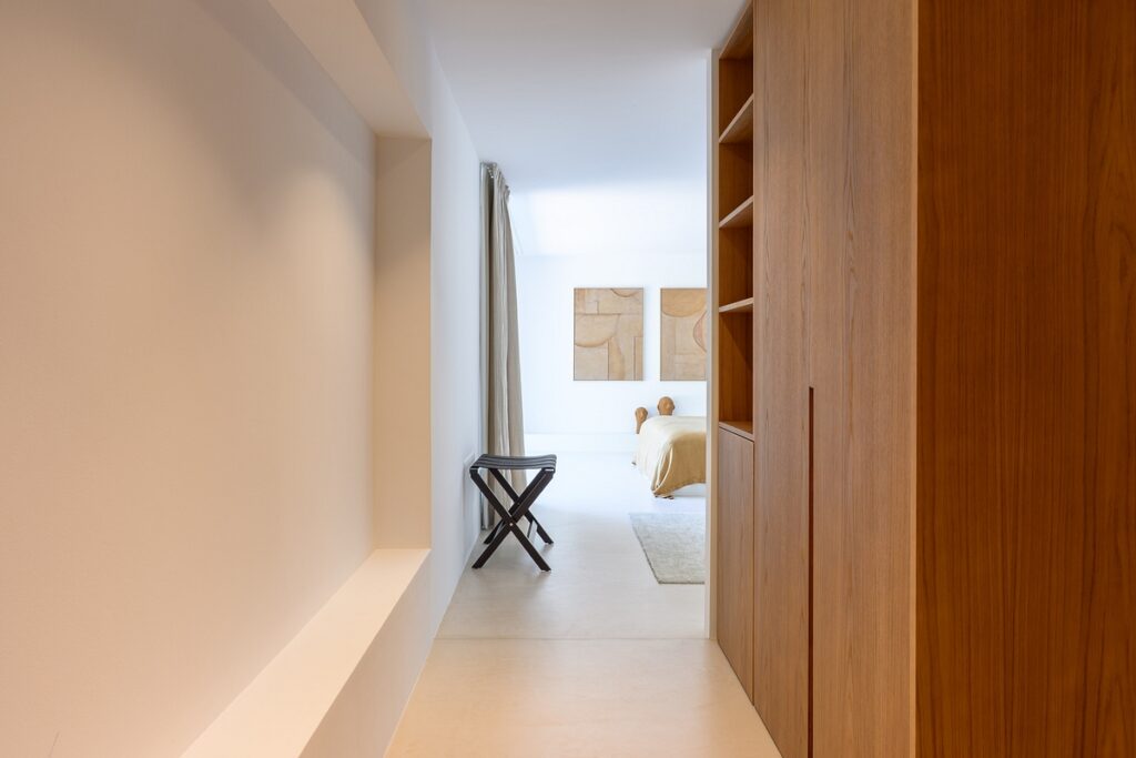 A minimalist hallway with light walls and wooden shelving leads to a bright bedroom featuring abstract art, a beige bed, a small stool, and floor-length curtains. David Pearson Travel
