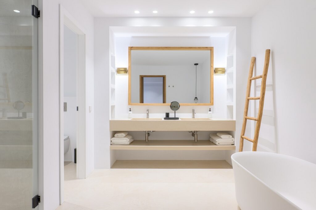 Modern bathroom with a large mirror above a double sink vanity, open shelves with towels, a ladder-style towel rack, a standalone bathtub, and minimal decor in a bright, white color scheme. David Pearson Travel