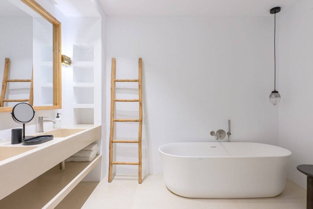 Minimalist bathroom with a freestanding white bathtub, wooden ladder-style towel rack, beige countertop with sink, large mirror with wooden frame, and a hanging pendant light against white walls. David Pearson Travel