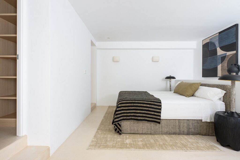 A modern minimalist bedroom with a bed featuring neutral bedding, a striped throw, two pillows, and a large abstract painting above the headboard. Two bedside tables with lamps and built-in shelving are visible. David Pearson Travel