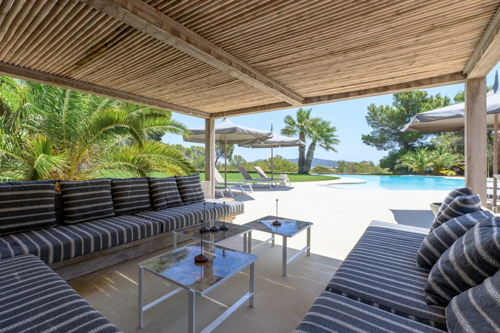 Outdoor lounge area with striped cushioned sofas under a wooden pergola, glass tables, palm trees, sun loungers, and a swimming pool with clear blue sky in the background. David Pearson Travel