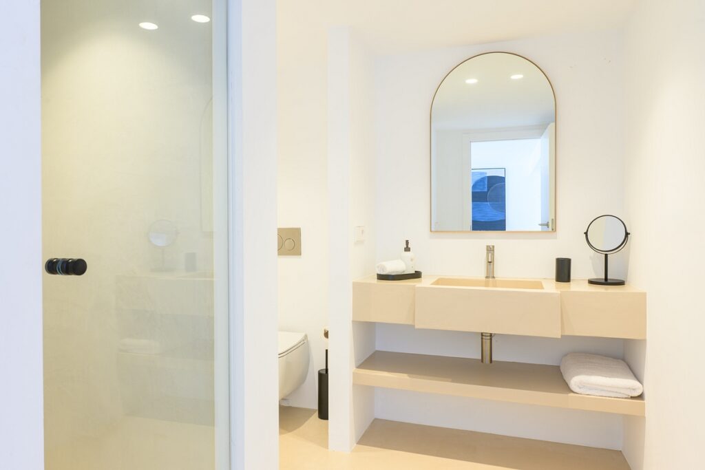 Modern, minimalistic bathroom with a beige vanity, large mirror, countertop sink, and open shelves with neatly folded towels. A glass shower and wall-mounted toilet are visible in a bright, clean space. David Pearson Travel