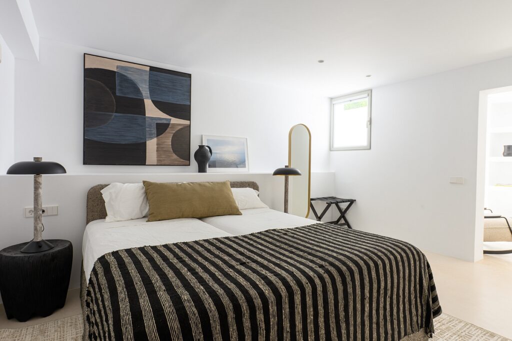 A modern bedroom with a double bed covered by a black and beige striped blanket, two black bedside tables with lamps, abstract wall art above the bed, a cushion, a full-length mirror, and a small window. David Pearson Travel