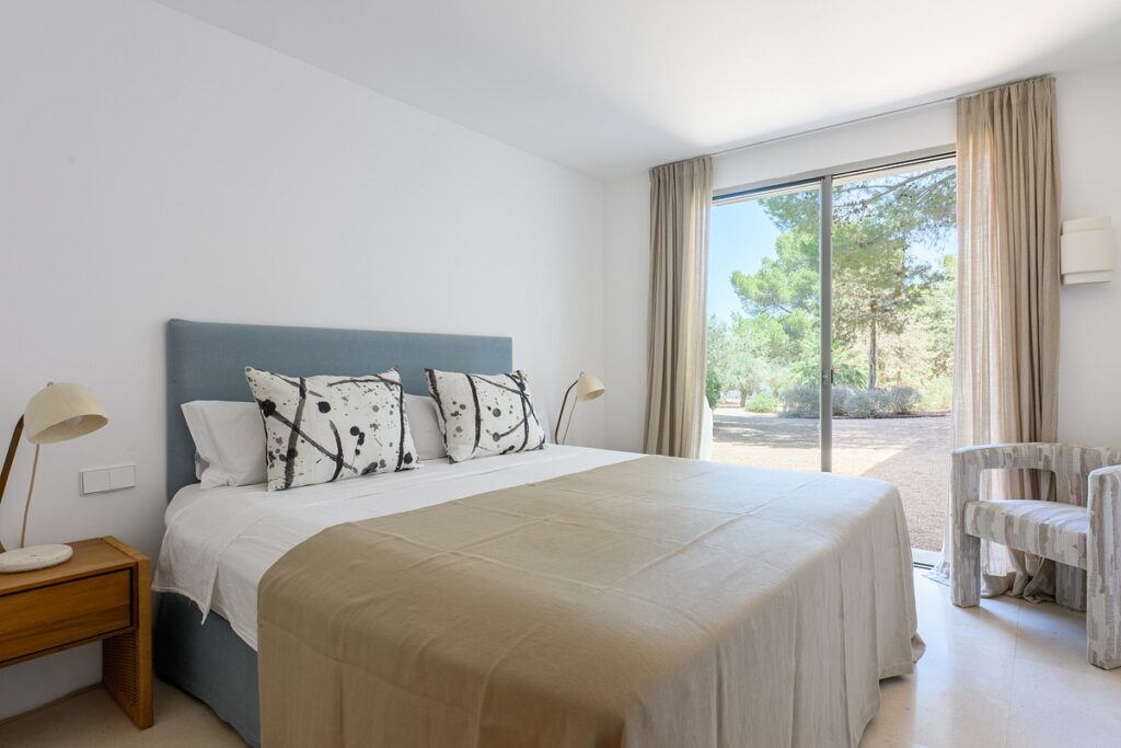 A bright, minimalist bedroom with a large bed, white bedding, beige throw, and patterned pillows. A bedside table with a lamp and a chair are beside the bed. Sliding glass doors open to a view of trees outside. David Pearson Travel