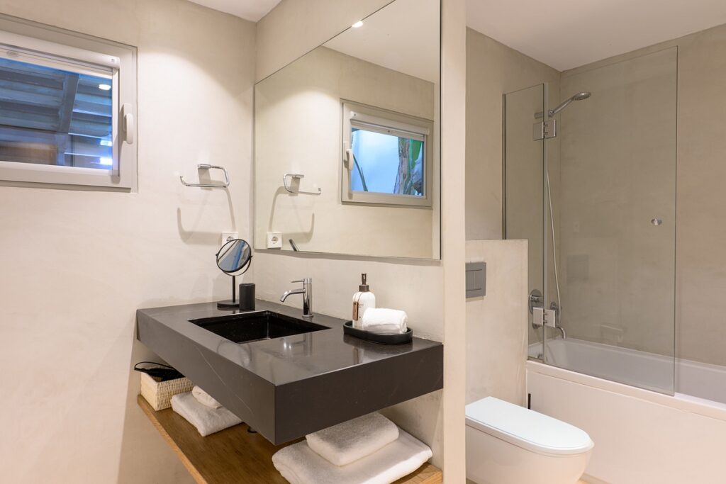 Modern bathroom with a large mirror, black sink, towels on a wooden shelf, wall-mounted toilet, and a bathtub with a glass shower screen. There is a small window and minimalistic decor. David Pearson Travel