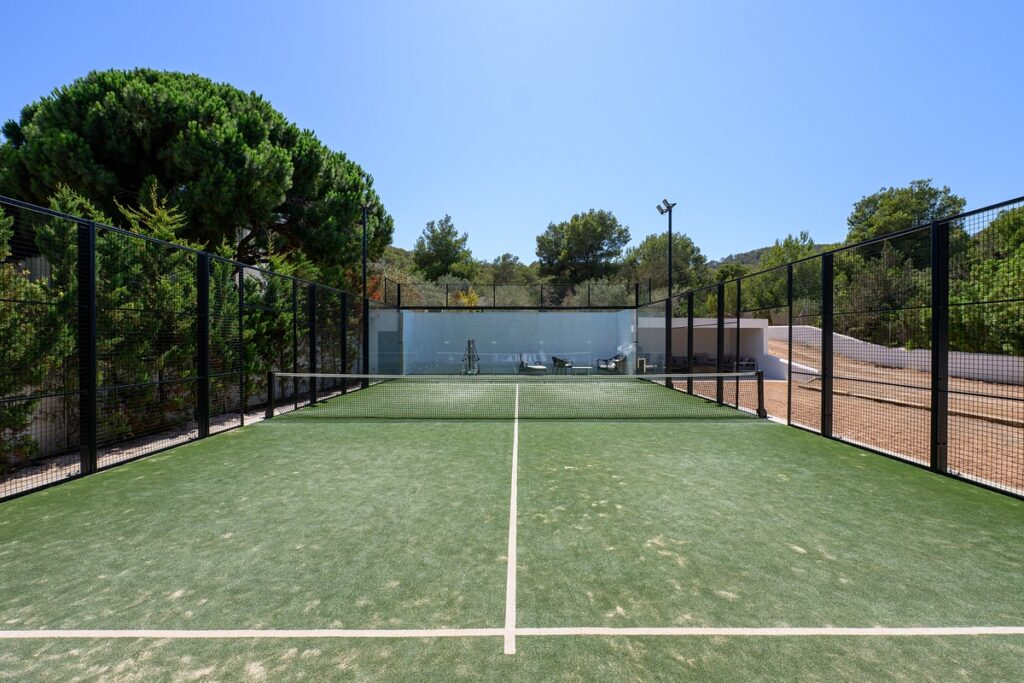 A green outdoor tennis or paddle court is surrounded by black fencing, with trees and shrubbery in the background under a clear blue sky. The court is empty and well-maintained. David Pearson Travel