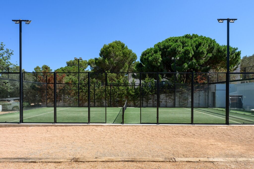 A fenced padel court with a net in the center, surrounded by trees and outdoor lights under a clear blue sky. The ground outside the court is covered in gravel. David Pearson Travel