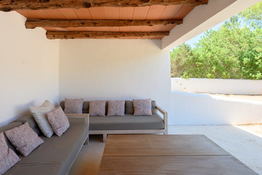 Outdoor patio with white walls, wooden ceiling beams, and two gray sofas with multiple cushions around a large wooden coffee table; greenery is visible outside the open wall. David Pearson Travel