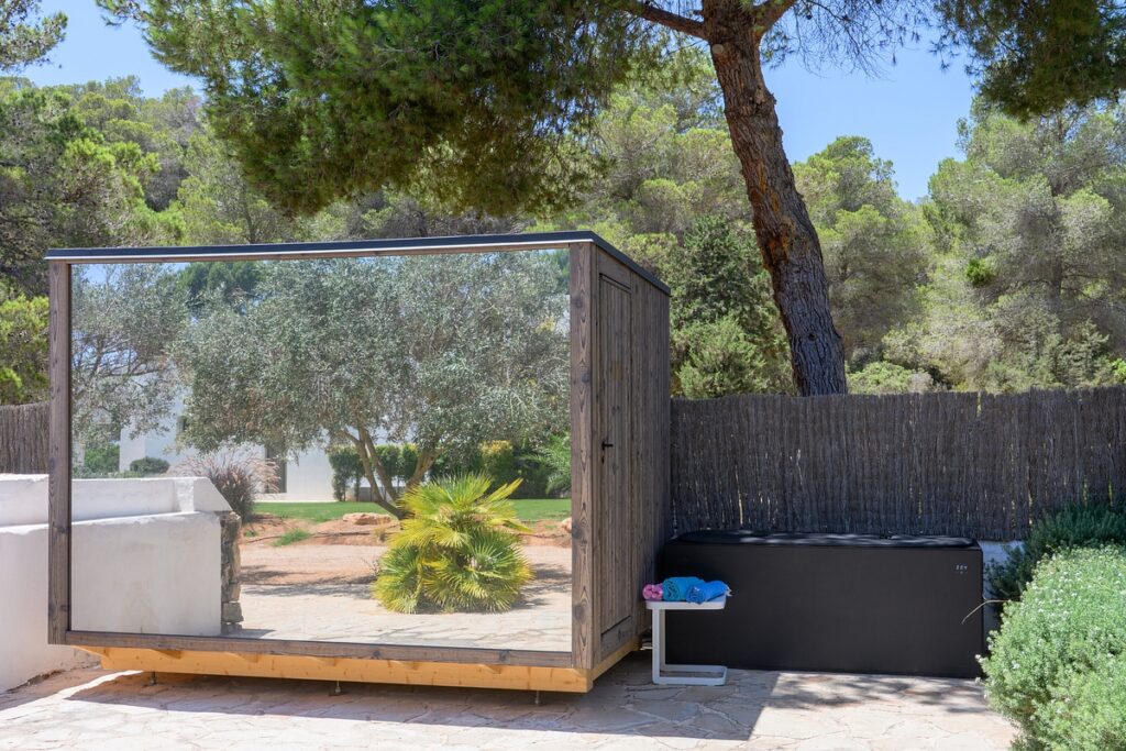 A small modern shed with reflective glass walls stands on a stone patio near trees. Next to it is a black storage box with towels on top, and a woven fence lines the background. David Pearson Travel