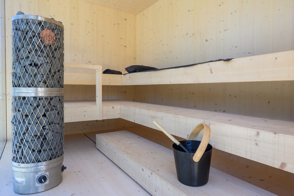 A modern wooden sauna with two-tiered benches, a tall metal sauna heater filled with rocks on the left, and a black bucket with a ladle on the floor. David Pearson Travel