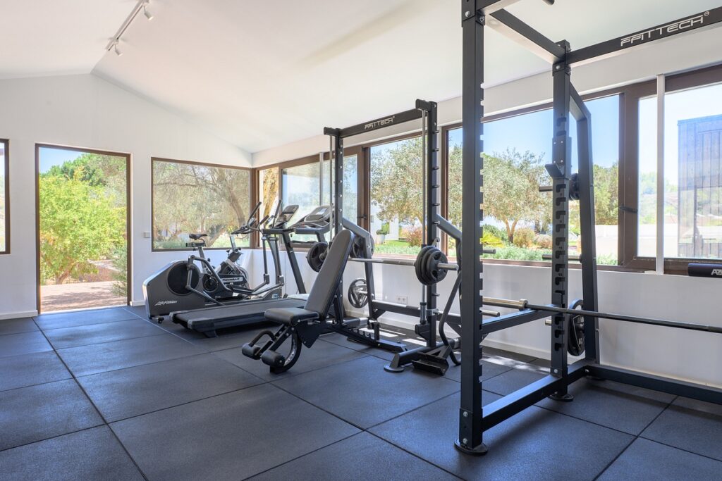 A bright, modern gym with large windows, featuring a squat rack, bench, stationary bike, elliptical machines, and rubber flooring. Greenery is visible outside. David Pearson Travel