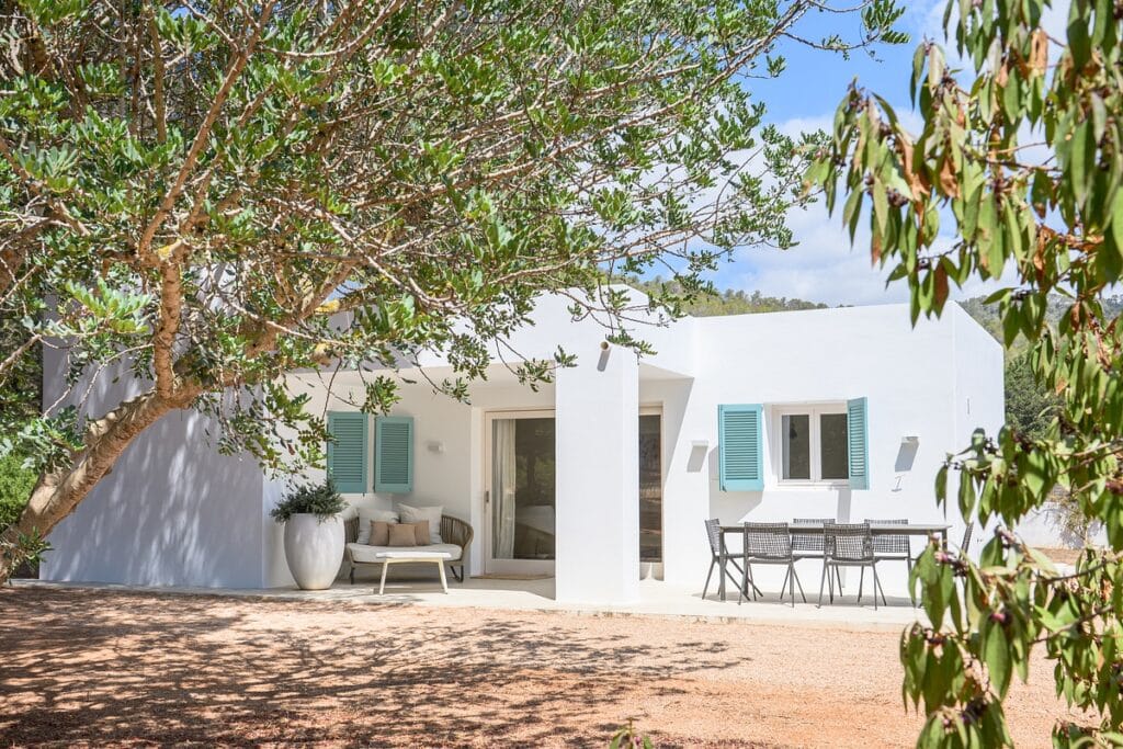 A modern white house with light blue shutters, a shaded outdoor dining area, large potted plant, and surrounding trees under a bright sky. David Pearson Travel