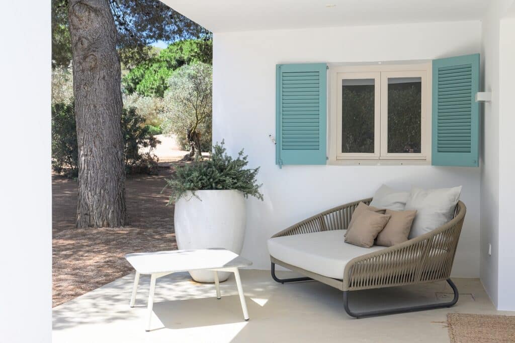 A cozy outdoor patio with a cushioned lounge chair, beige and white pillows, a small white table, a large potted plant, and a white wall with a window featuring blue-green shutters. Pine trees are visible in the background. David Pearson Travel
