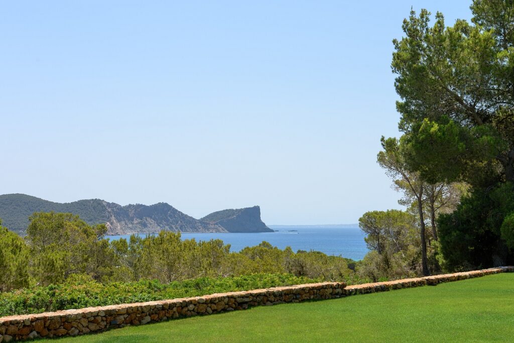 A scenic view of a lush green lawn bordered by a stone wall, with trees on the right and hills leading down to a calm blue sea under a clear sky in the background. David Pearson Travel