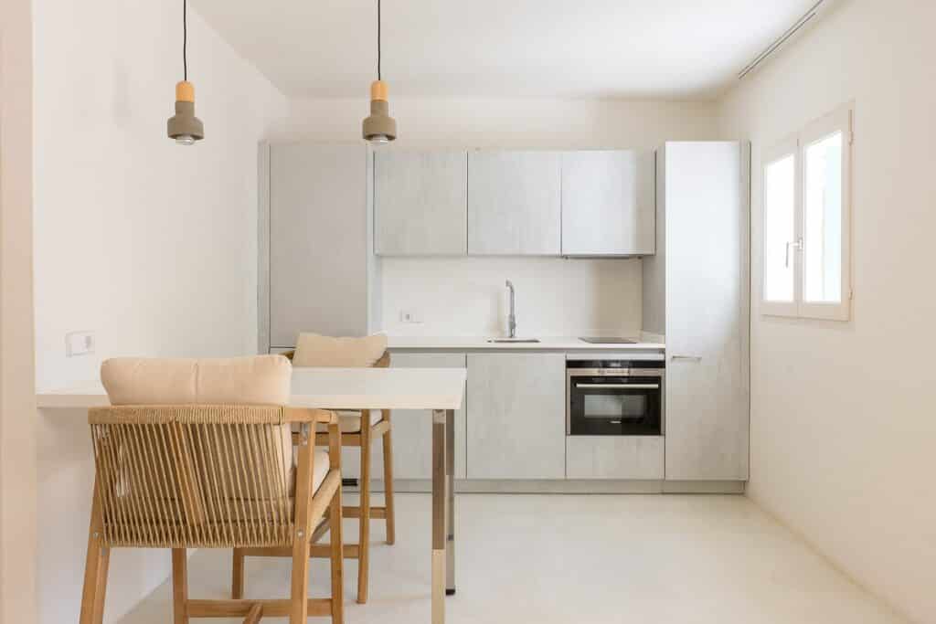 Minimalist kitchen with light grey cabinets, built-in oven, cooktop, and sink. Two wooden chairs with cushions are placed at a white table. Pendant lights hang from the ceiling, and natural light enters through a window. David Pearson Travel