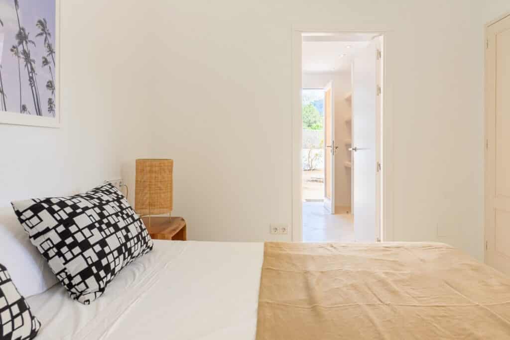 Minimalist bedroom with a bed topped with geometric-patterned pillows and a beige blanket, a wooden nightstand with a wicker lamp, and a door open to a bright hallway with natural light. David Pearson Travel