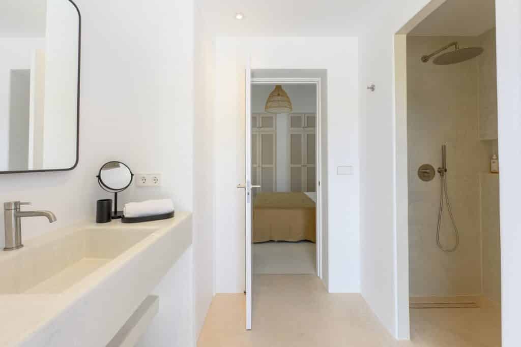 Modern, minimalist bathroom with a beige sink, mirror, and toiletries on the left, and an open door revealing a bedroom. On the right, there is a walk-in shower with a rain showerhead. David Pearson Travel