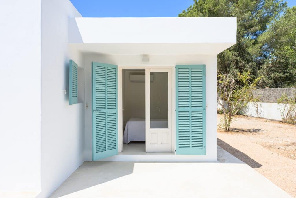 A white building with open light blue shutters and a glass door, revealing a simple bedroom inside. The exterior is minimalistic, with a clear sky and some trees in the background. David Pearson Travel