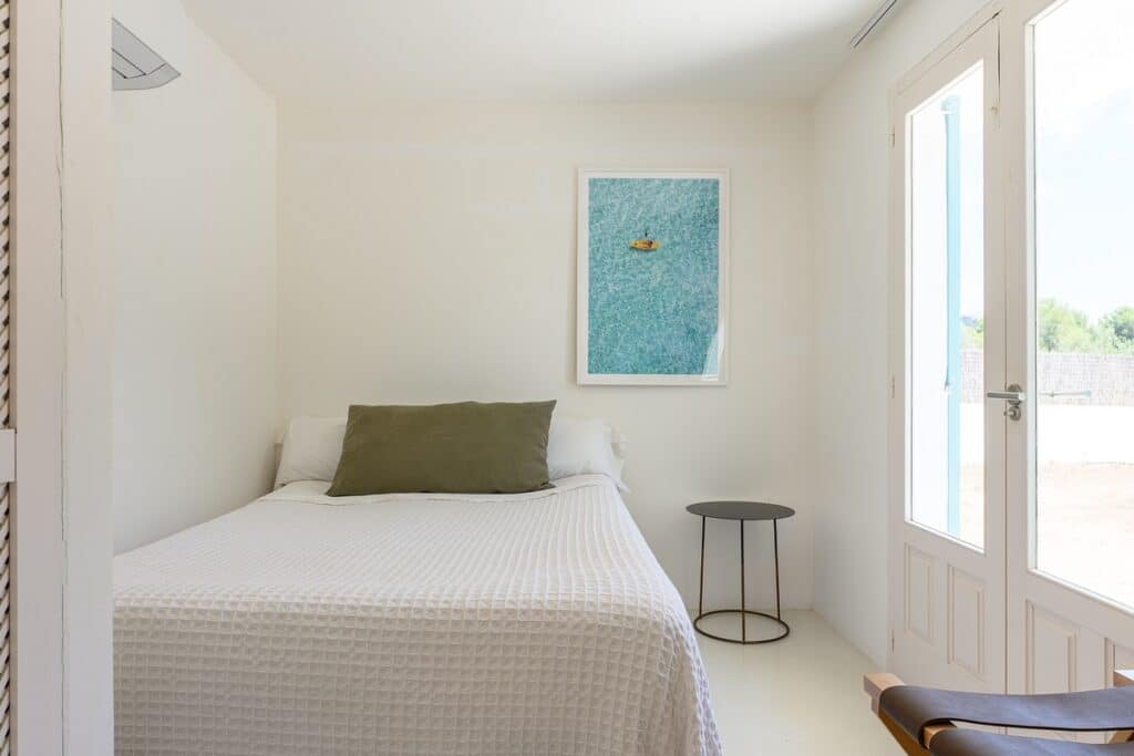 A minimalist bedroom with a white bed, a green pillow, a small round black side table, a framed picture on the wall, and glass doors letting in natural light. David Pearson Travel