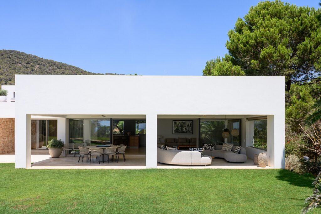Modern white villa with large open patios, outdoor dining and seating areas, surrounded by green lawn, trees, and hills under a clear blue sky. David Pearson Travel