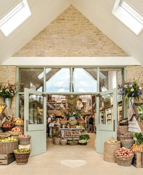 A rustic farm shop in The Cotswolds with stone walls, an open double door, baskets of fresh fruits and vegetables, and shelves of goods inside. Skylights and floral arrangements add a bright, welcoming atmosphere. David Pearson Travel