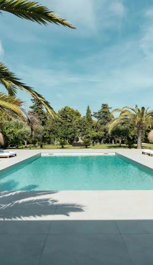 villa-maggie-22.jpg - Edited A rectangular outdoor pool surrounded by white lounge chairs and straw parasols, featured on the Home Page, with palm trees and greenery in the background under a blue sky. David Pearson Travel
