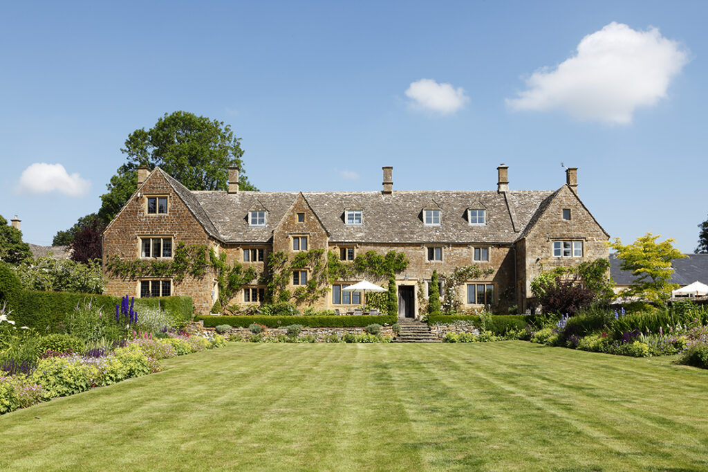 Large stone country house with steep roofs and multiple chimneys, surrounded by lush gardens and a neatly mown lawn, under a bright blue sky with a few clouds. David Pearson Travel