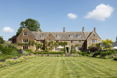 1 Large stone country house with steep roofs and multiple chimneys, surrounded by lush gardens and a neatly mown lawn, under a bright blue sky with a few clouds. David Pearson Travel