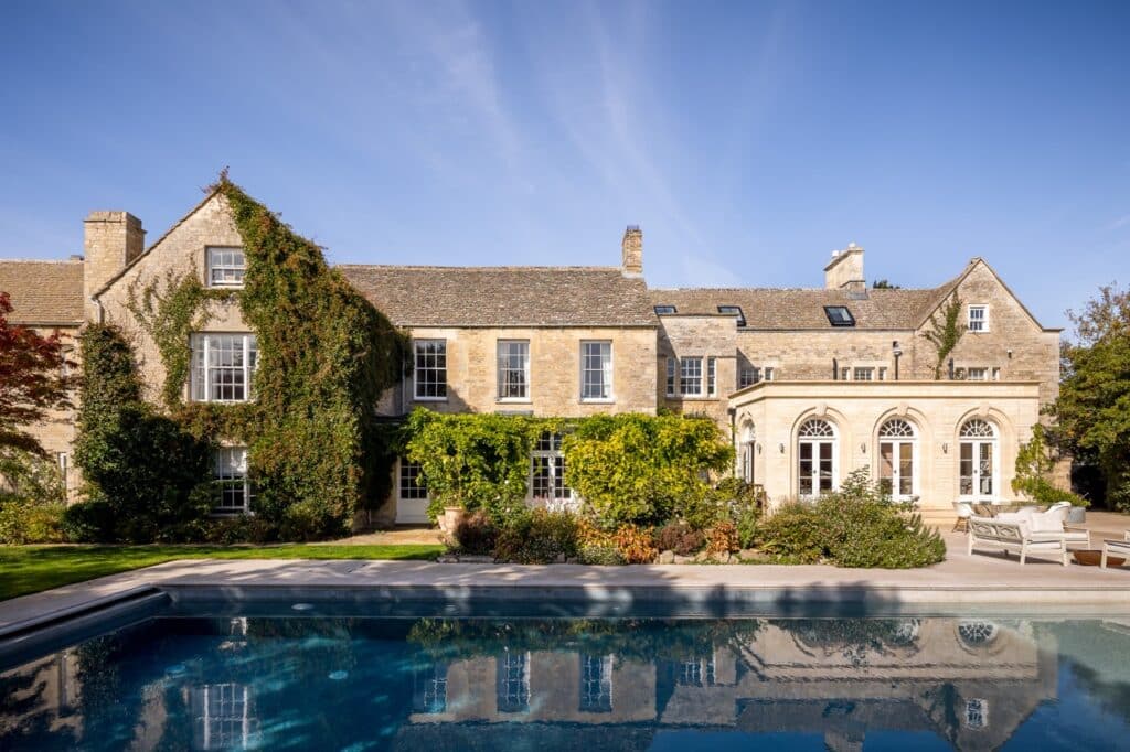 A large stone country house with ivy climbing the walls, tall windows, and arched doorways. In front is a clear blue swimming pool surrounded by a patio, outdoor furniture, and lush landscaped gardens. David Pearson Travel