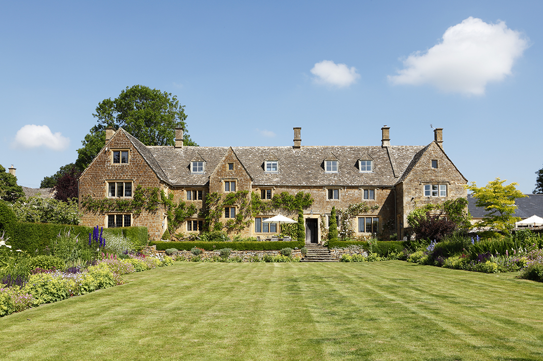 Large stone country house with steep roofs and multiple chimneys, surrounded by lush gardens and a neatly mown lawn, under a bright blue sky with a few clouds. David Pearson Travel