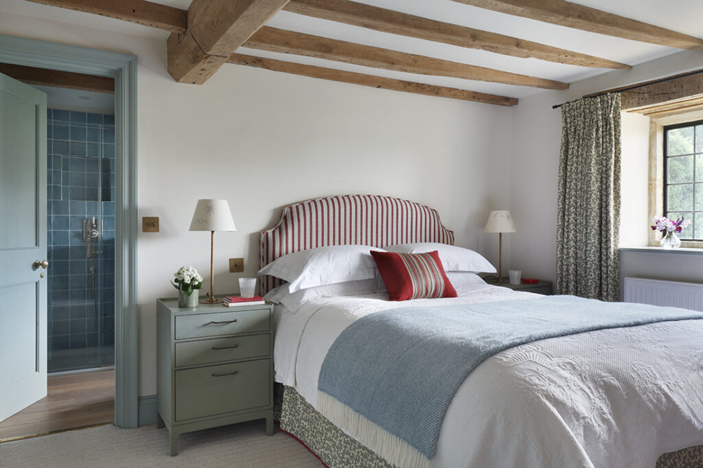A cosy bedroom with exposed wooden ceiling beams, a striped upholstered headboard, a neatly made bed with layered pillows and blankets, two green bedside tables with lamps, and an adjoining bathroom with blue tiles. David Pearson Travel