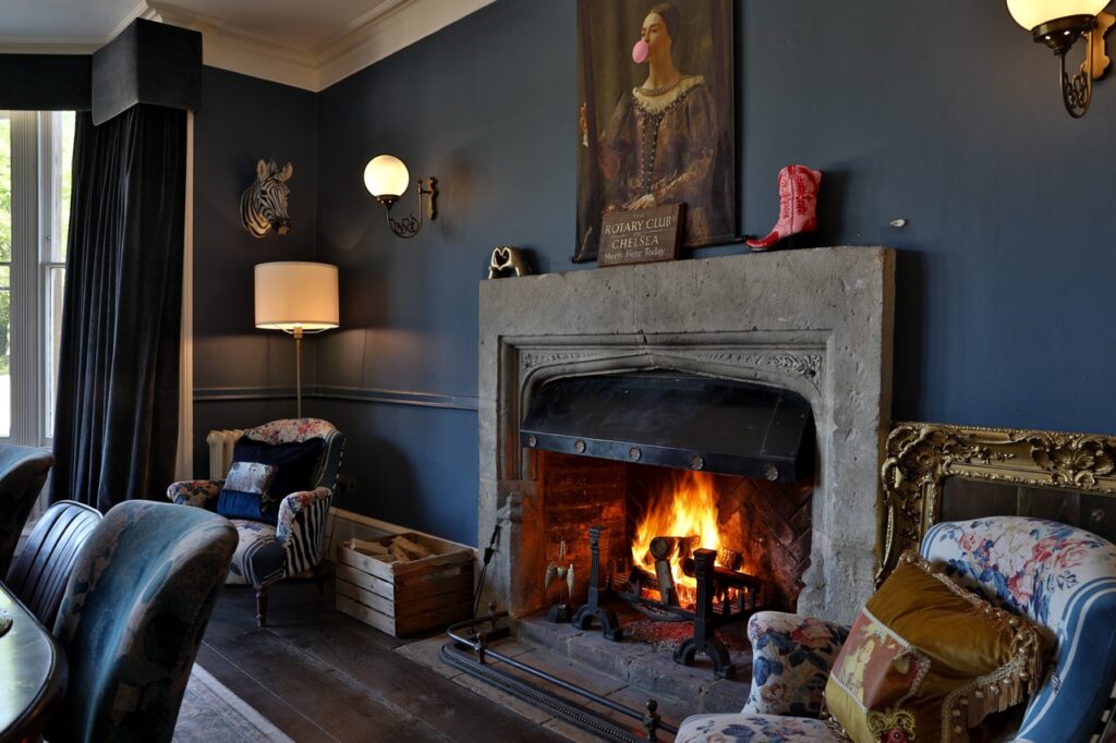 A cosy living room with a lit fireplace, blue walls, vintage armchairs, a standard lamp, a large window with curtains, and quirky decor including a zebra head and a painting of a woman blowing bubble gum. David Pearson Travel