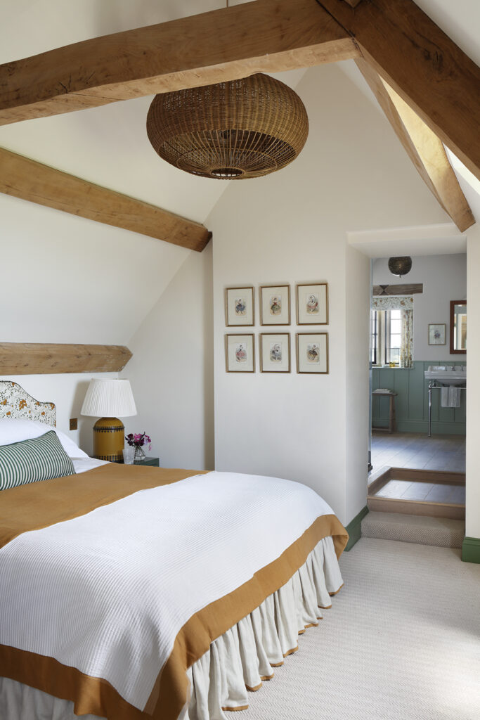A cosy bedroom with exposed wooden beams, a double bed with white and mustard bedding, a wicker ceiling light, framed art on the wall, and a view into a bathroom with green cupboards. David Pearson Travel