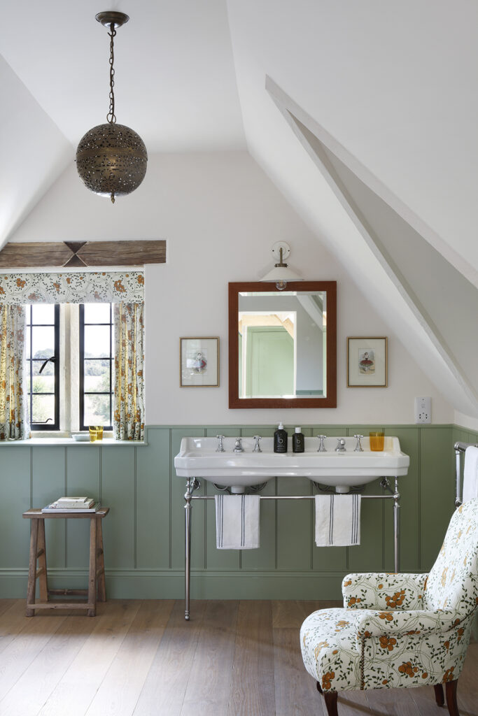 A cosy bathroom with light green wainscoting, a double basin, floral curtains, a floral armchair, a wooden stool, a round pendant light, and a mirror above the basin, set in an attic space with sloped ceilings. David Pearson Travel