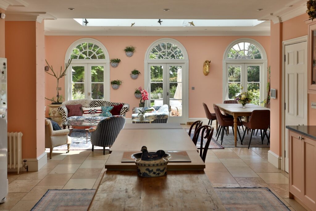 A spacious, sunlit kitchen and dining area with peach walls, arched windows, potted plants, a cosy seating area with patterned chairs, and a large wooden table on tiled floors. Light streams in through the windows. David Pearson Travel