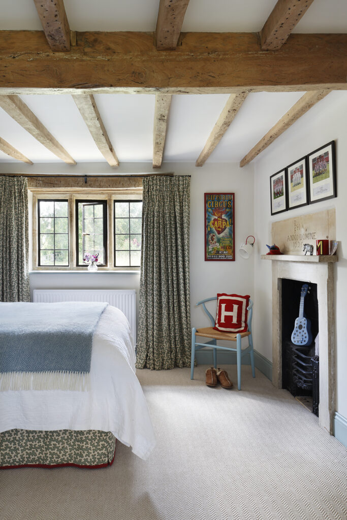 A cosy bedroom with exposed wooden ceiling beams, a bed with white and blue bedding, patterned curtains, a blue chair with a red H cushion, wall art, and a fireplace decorated with a blue toy guitar. David Pearson Travel