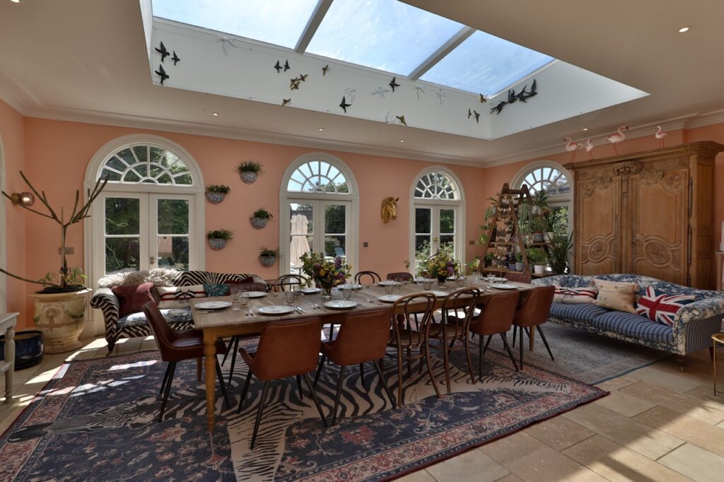 A bright dining room with a large skylight, pink walls, a long wooden table laid for a meal, eclectic chairs, a patterned rug, wall plants, a striped sofa, and decorative accents, including hanging birds and flamingos. David Pearson Travel