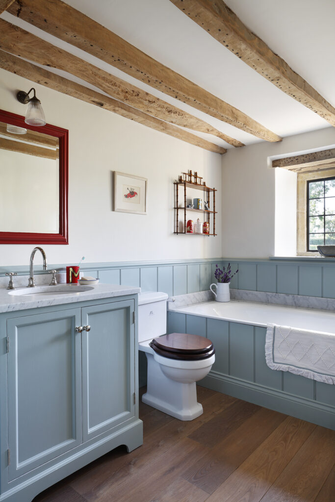 A rustic bathroom with wooden ceiling beams, light blue cabinetry, a white washbasin, toilet with dark wooden seat, bath, and a red-framed mirror. Sunlight enters through a window, creating a cosy, inviting atmosphere. David Pearson Travel