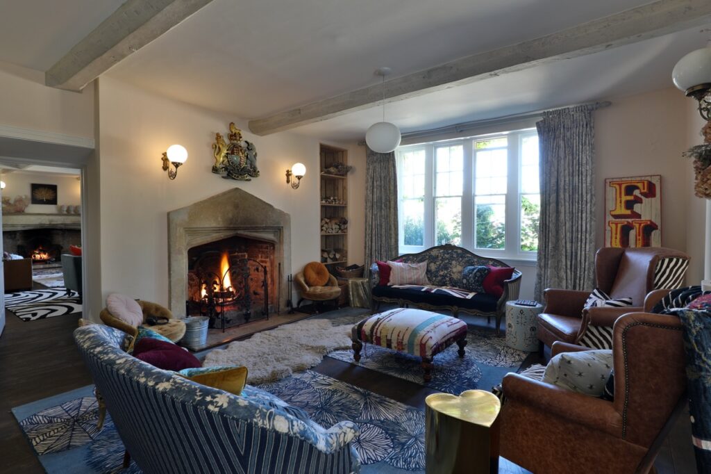Cosy living room with a lit fireplace, vintage furniture, patterned rugs, and large windows with curtains letting in natural light. Shelves hold decor items and books, and the space features a warm, eclectic atmosphere. David Pearson Travel
