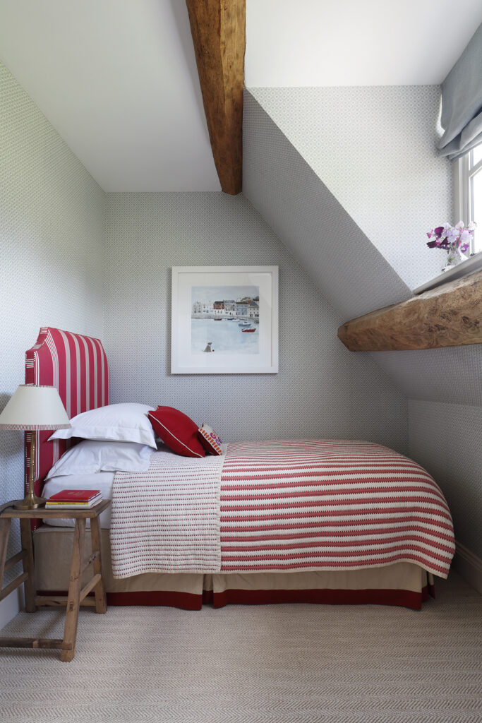 A cosy attic bedroom with a single bed featuring red and white striped bedding, a wooden stool as a bedside table, a framed artwork on the wall, and exposed wooden beams under a slanted ceiling. David Pearson Travel
