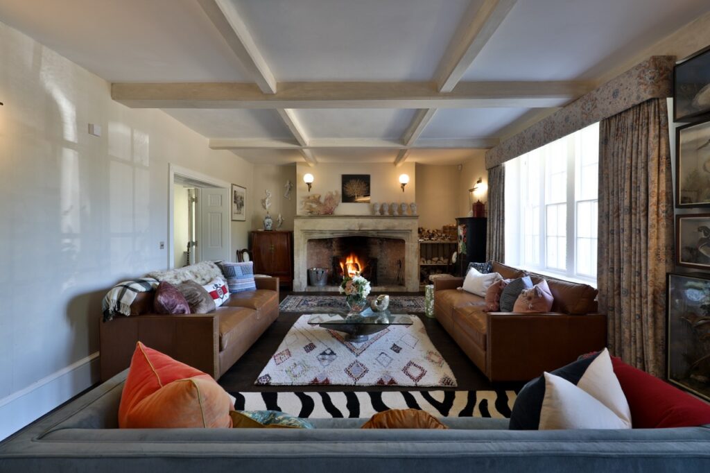 A cosy living room with a roaring fireplace, two brown leather sofas, a blue settee with colourful cushions, a patterned rug, large windows with curtains, and various decorative items on shelves and walls. David Pearson Travel