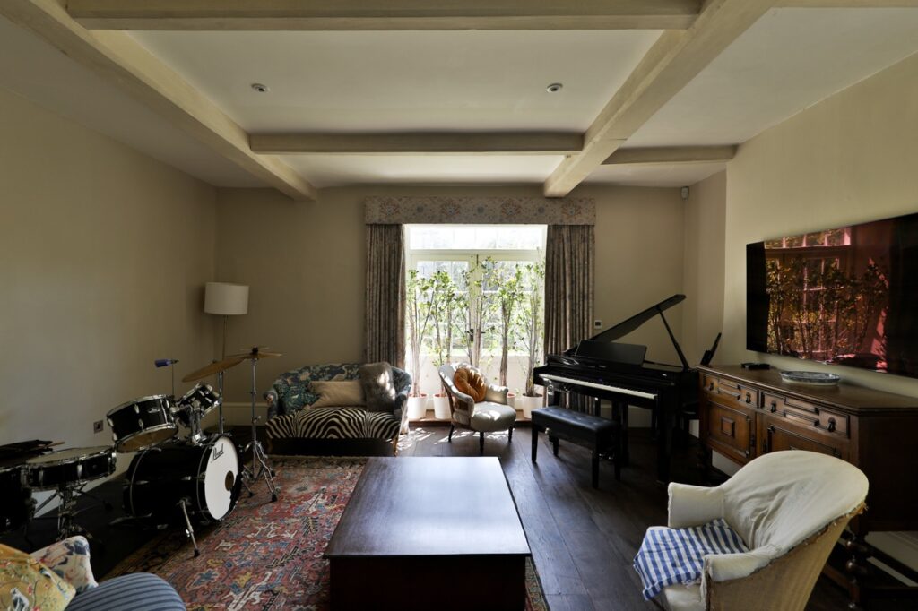 A cosy living room with a drum kit, grand piano, sofas, armchairs, a large TV on a wooden cabinet, a coffee table, and a large window with plants outside. Natural light fills the room. David Pearson Travel
