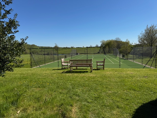 Three wooden benches face a fenced tennis court on a sunny day, with green grass in the foreground and open countryside and blue sky in the background. David Pearson Travel