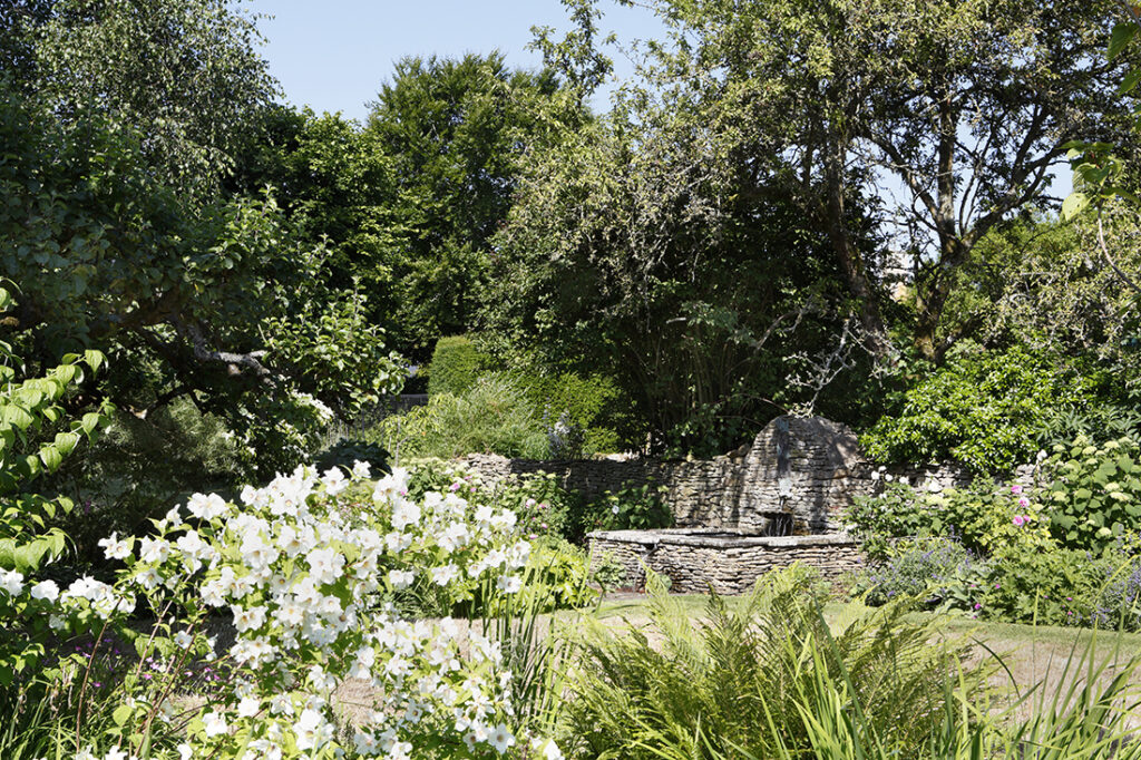 A lush garden with green trees and bushes, white flowers in the foreground, and a stone wall with a small fountain in the background under a clear blue sky. David Pearson Travel