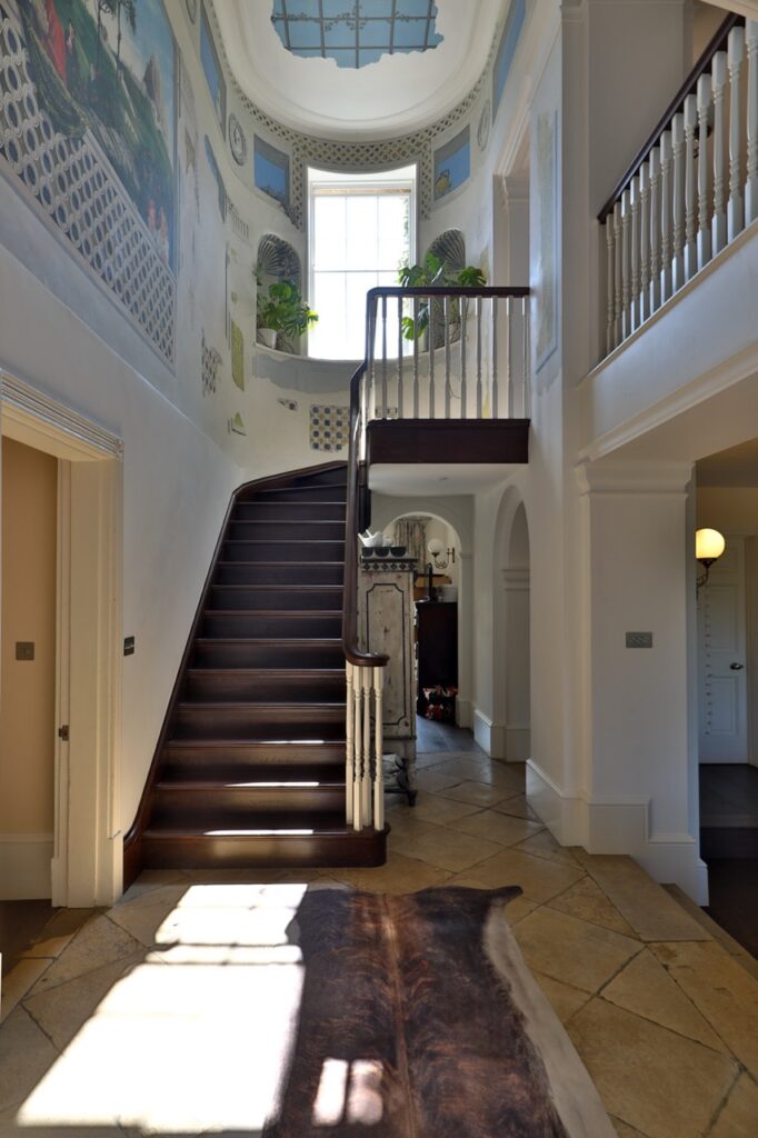 A grand, sunlit hallway with a curved wooden staircase, white bannisters, murals on the walls, a large window above, and a skylight. Light streams onto the tiled floor, highlighting a brown rug. David Pearson Travel